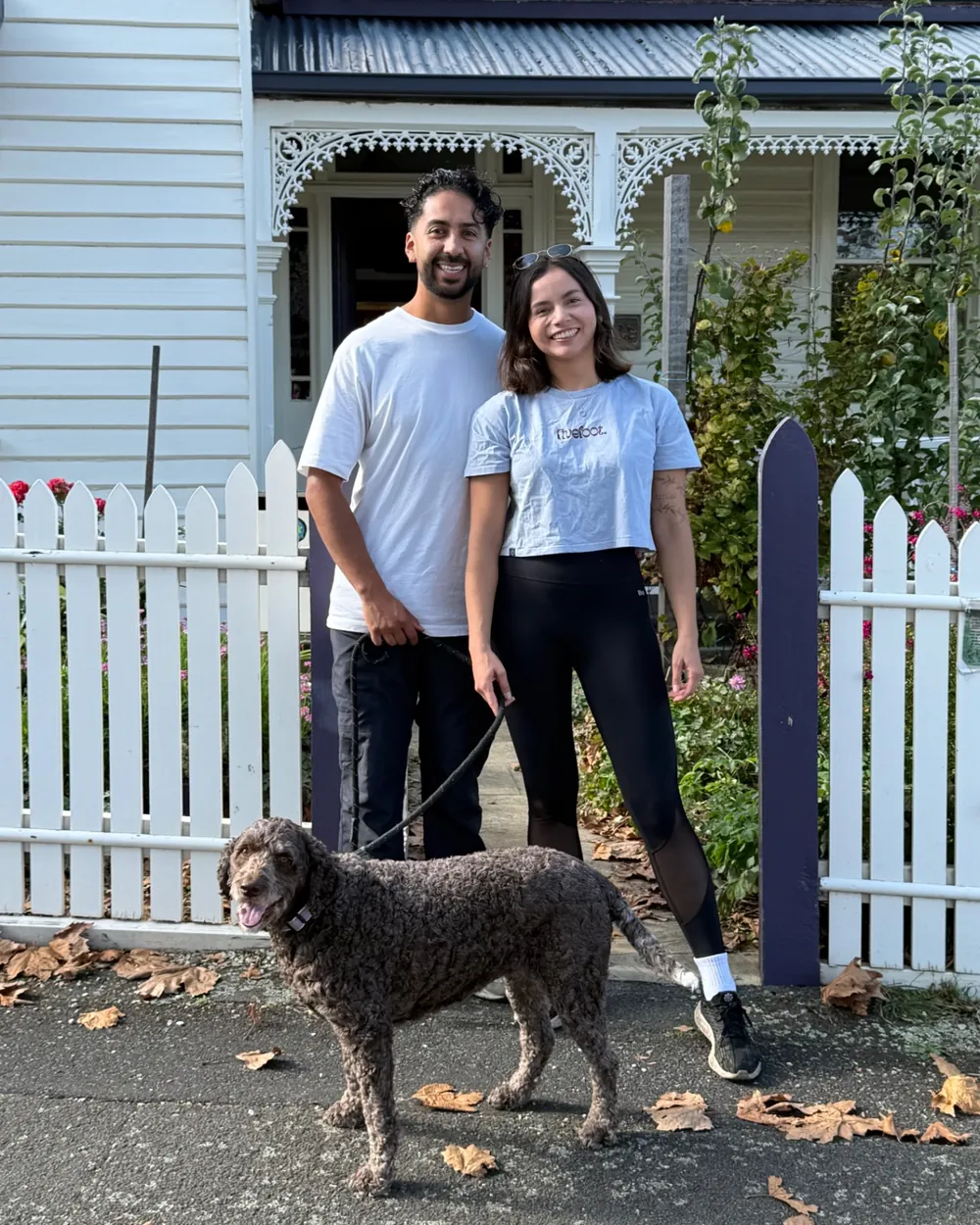Yen and Nico standing side by side in front of a white weatherboard house with their curly-coated dog on the path
