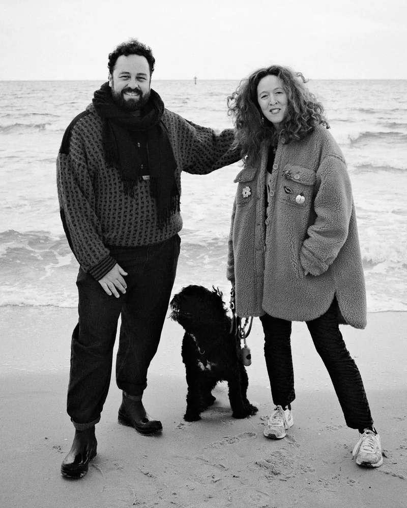 A black and white photo of Ellen and Ede standing close together on a beach with their Portuguese Water Dog Tilde between them on the sand