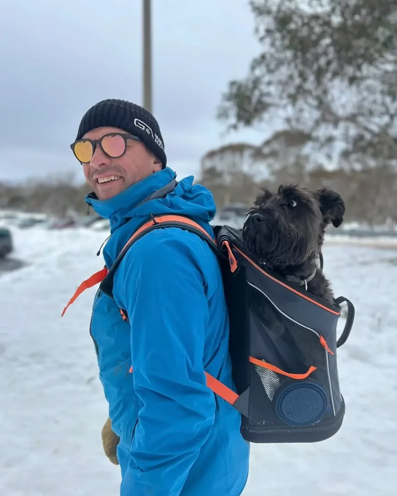 A man in a blue jacket and beanie standing in the snow with his dog Hunzo looking out of the carrier backpack on his back